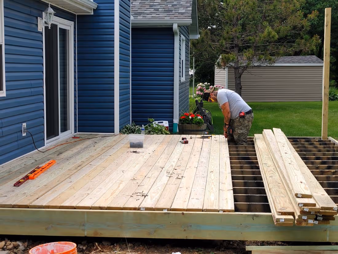 Man working on building a wooden deck attached to a blue house in a green backyard.
