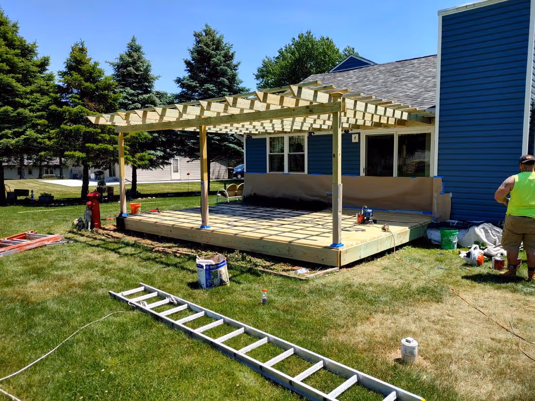 Construction of a wooden pergola frame on a raised deck attached to a blue house, with tools and a worker nearby on green grass.
