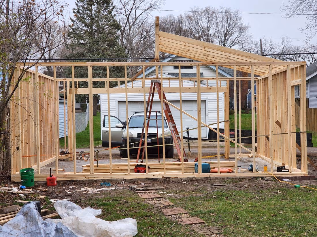 Wooden framework of a small building under construction with a ladder and a parked car in the background.
