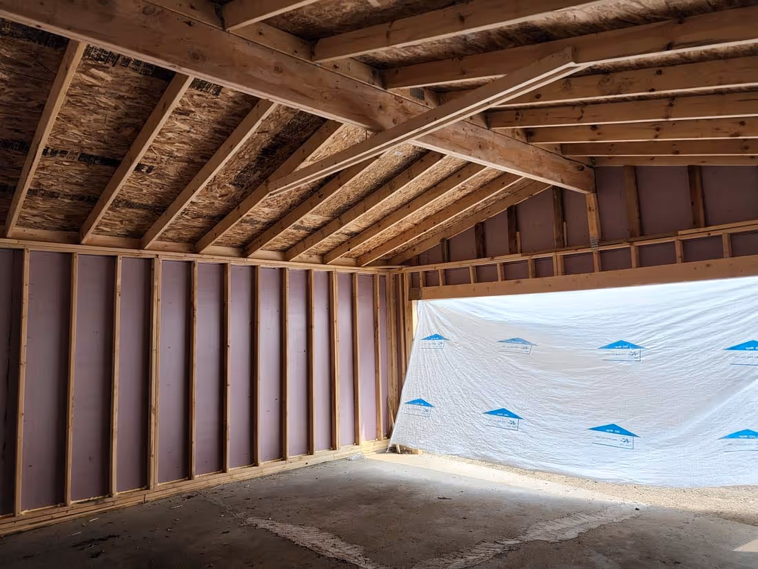 Interior of an unfinished wooden garage with exposed beams and a large opening covered by a white plastic sheet.