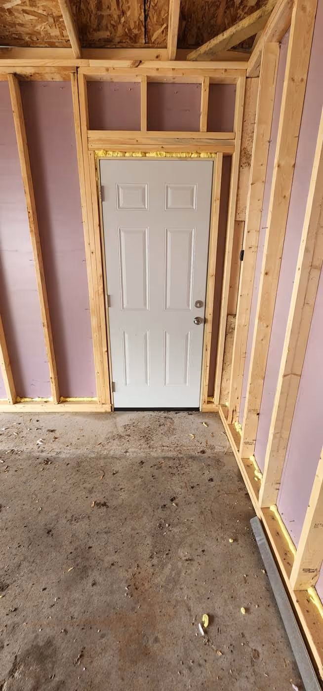 White panel door installed in an unfinished wooden frame wall with pink insulation panels and a concrete floor.