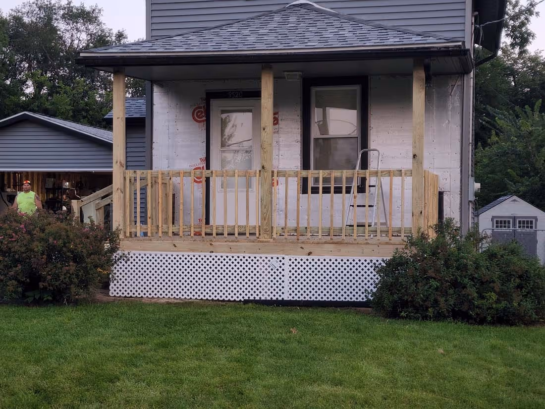Front porch of a small house under construction with wooden railing, a white door, a window, and green lawn in the foreground.