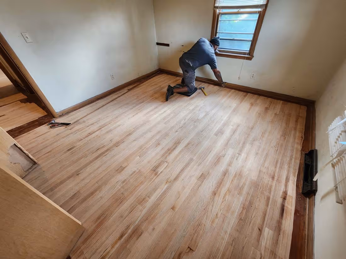 Worker installing or refinishing hardwood floor in an empty room with beige walls and a window.