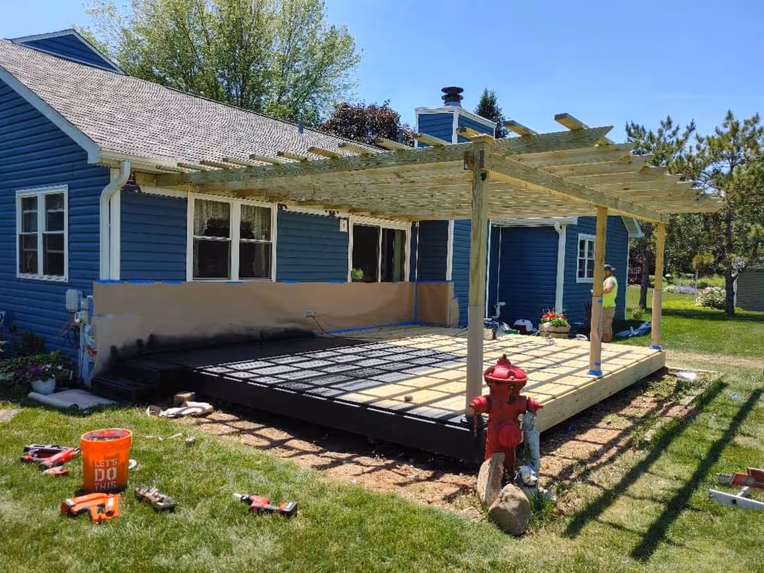 Wooden patio deck under construction attached to a blue house with a pergola overhead and tools scattered on the lawn.
