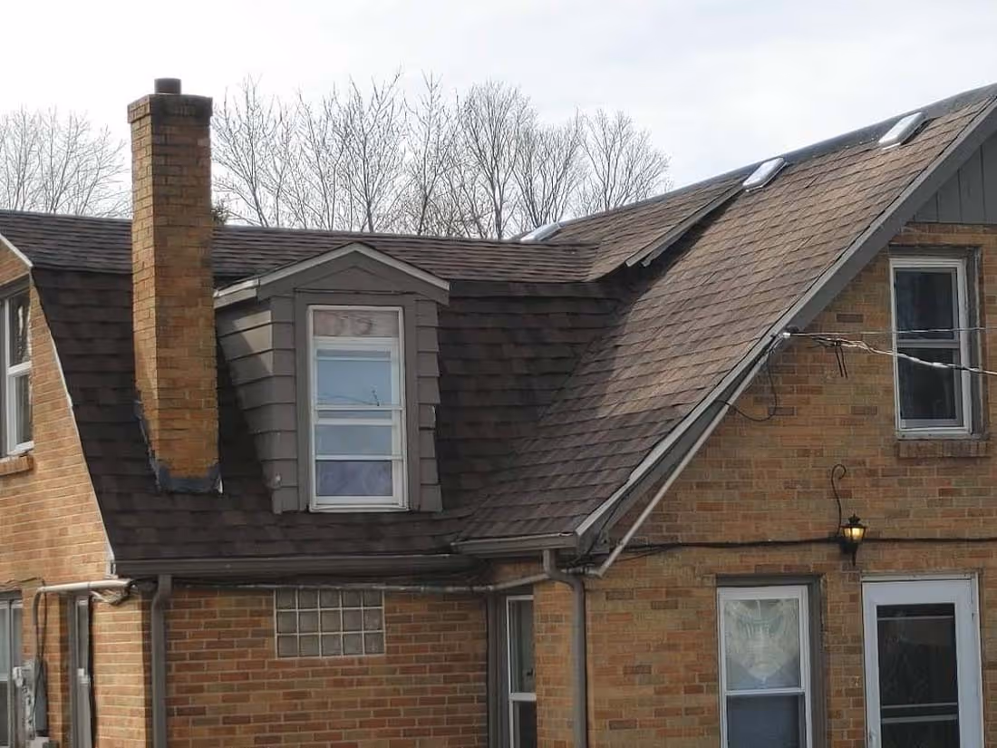 Brick house with multiple windows, a tall chimney, and a brown shingle roof with skylights and a dormer window.