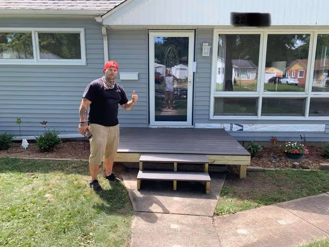 Man with tattoos wearing a red cap and black shirt giving a thumbs up while standing next to a newly built small wooden front porch with two steps in front of a gray house.