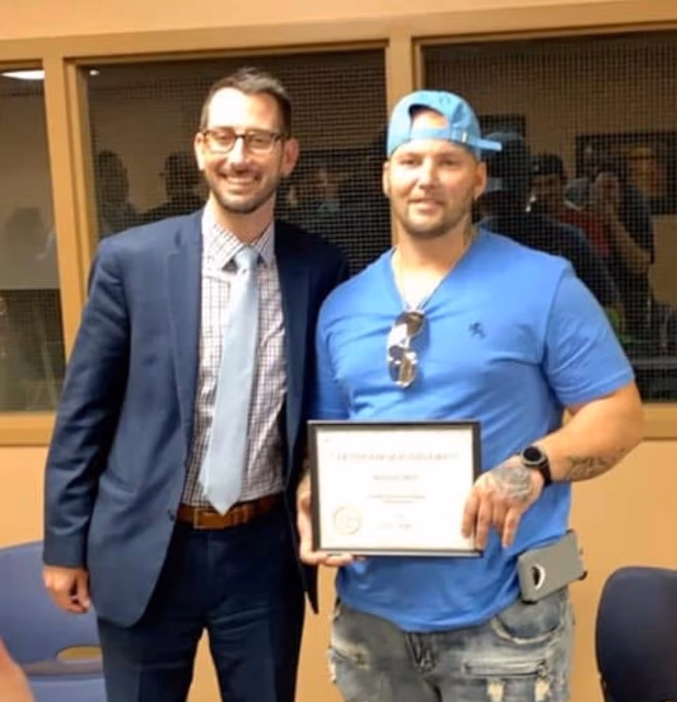 Two men standing together, one in a suit and tie smiling, the other in a blue shirt and backward cap holding a framed certificate.