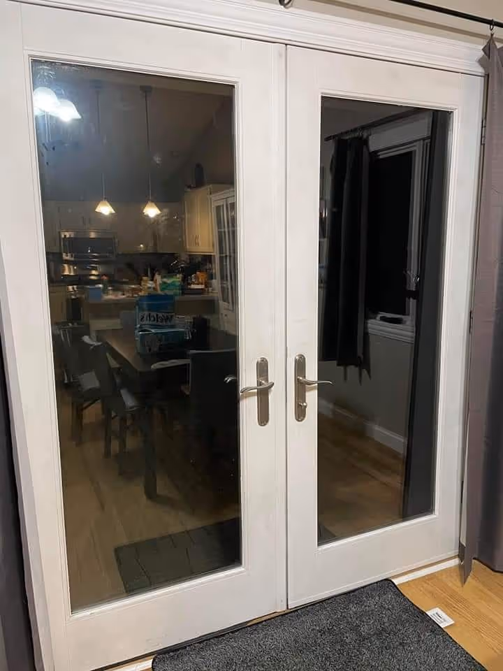 White-framed double glass doors with silver handles showing a dimly lit kitchen and dining area inside.