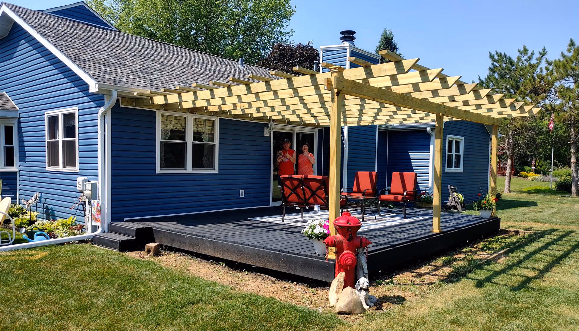 Blue house with a wooden pergola over a black deck furnished with red cushioned chairs and a red fire hydrant with pet statues in front.