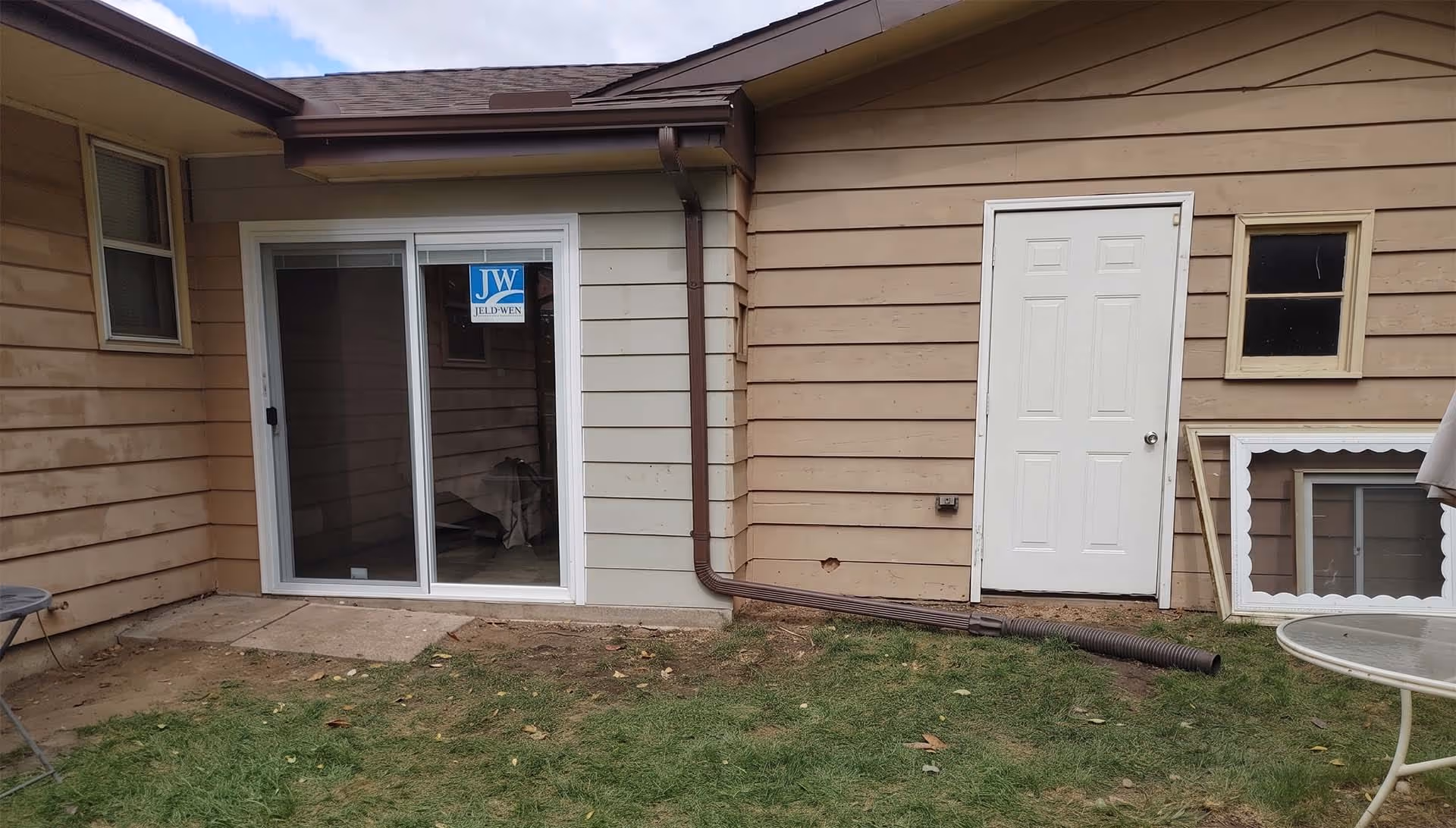 Backyard view of a house showing a white sliding glass door with a Jeld-Wen sticker, a white single door, a small window, and a brown downspout drainage pipe extending onto the grass.