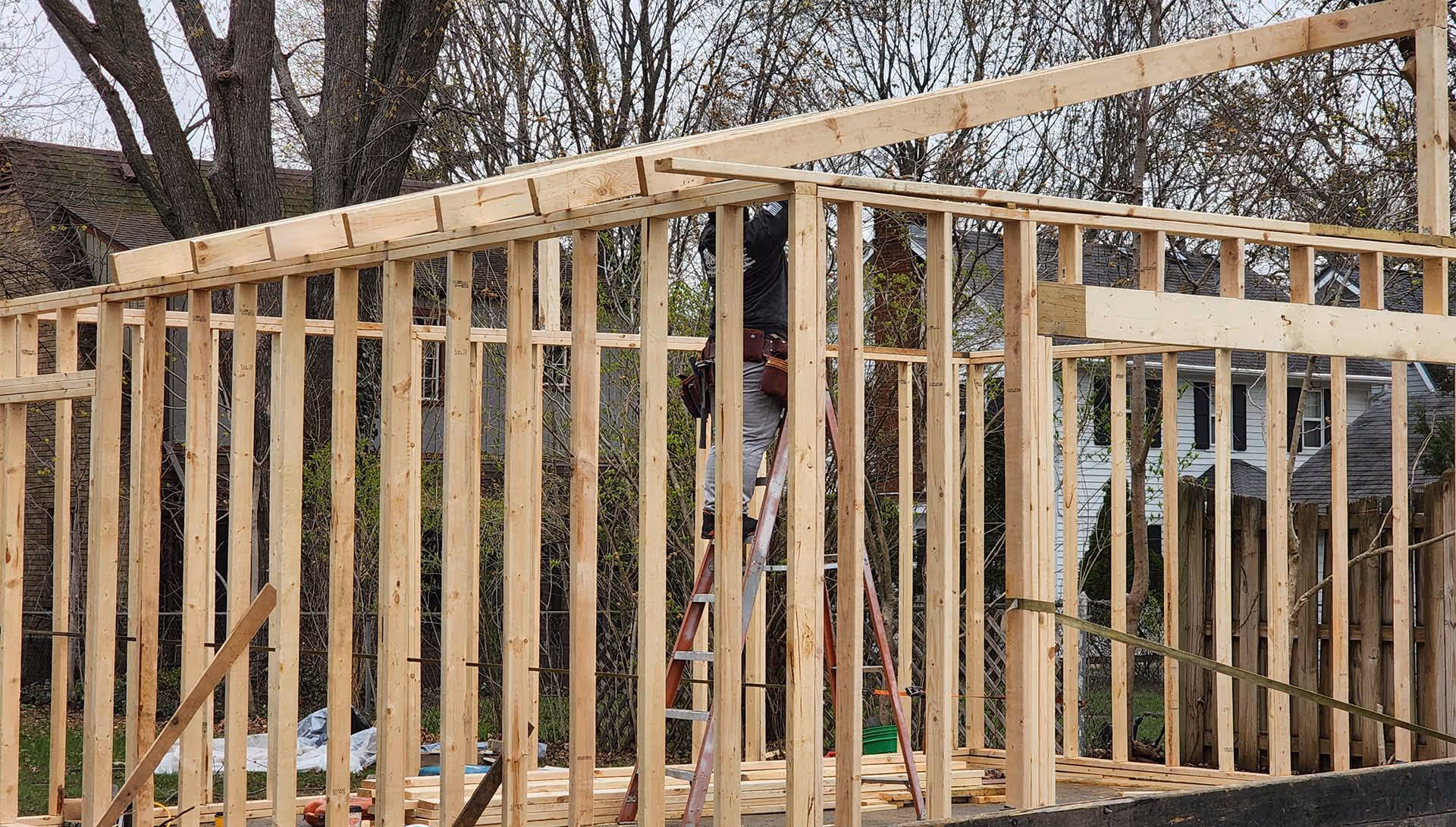 Construction worker on a ladder building wooden frame of a house outdoors in a backyard.
