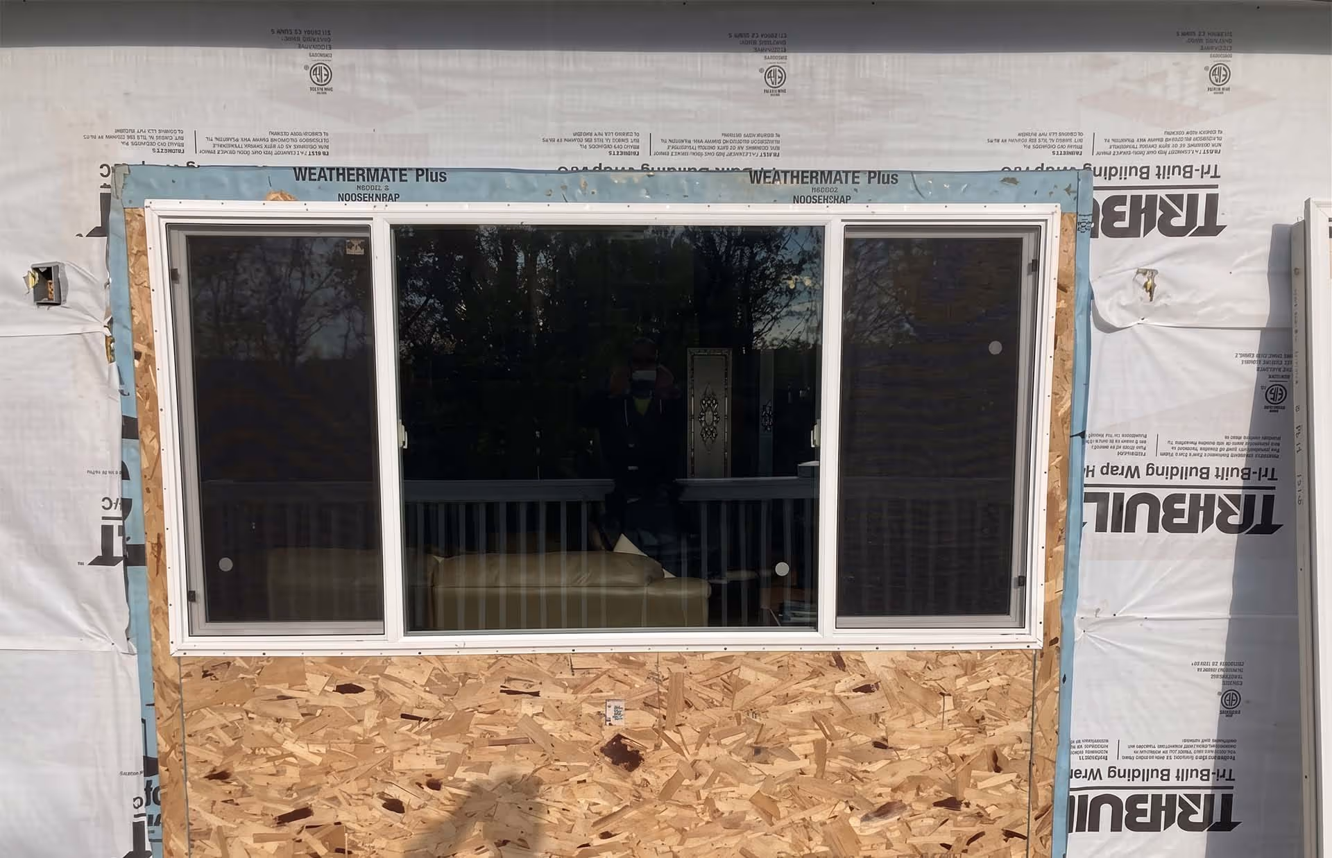 Three-panel window installed on plywood sheathing, part of a building under construction with house wrap visible.