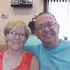Smiling elderly man in turquoise shirt sitting next to a smiling elderly woman in red top with glasses, indoors.