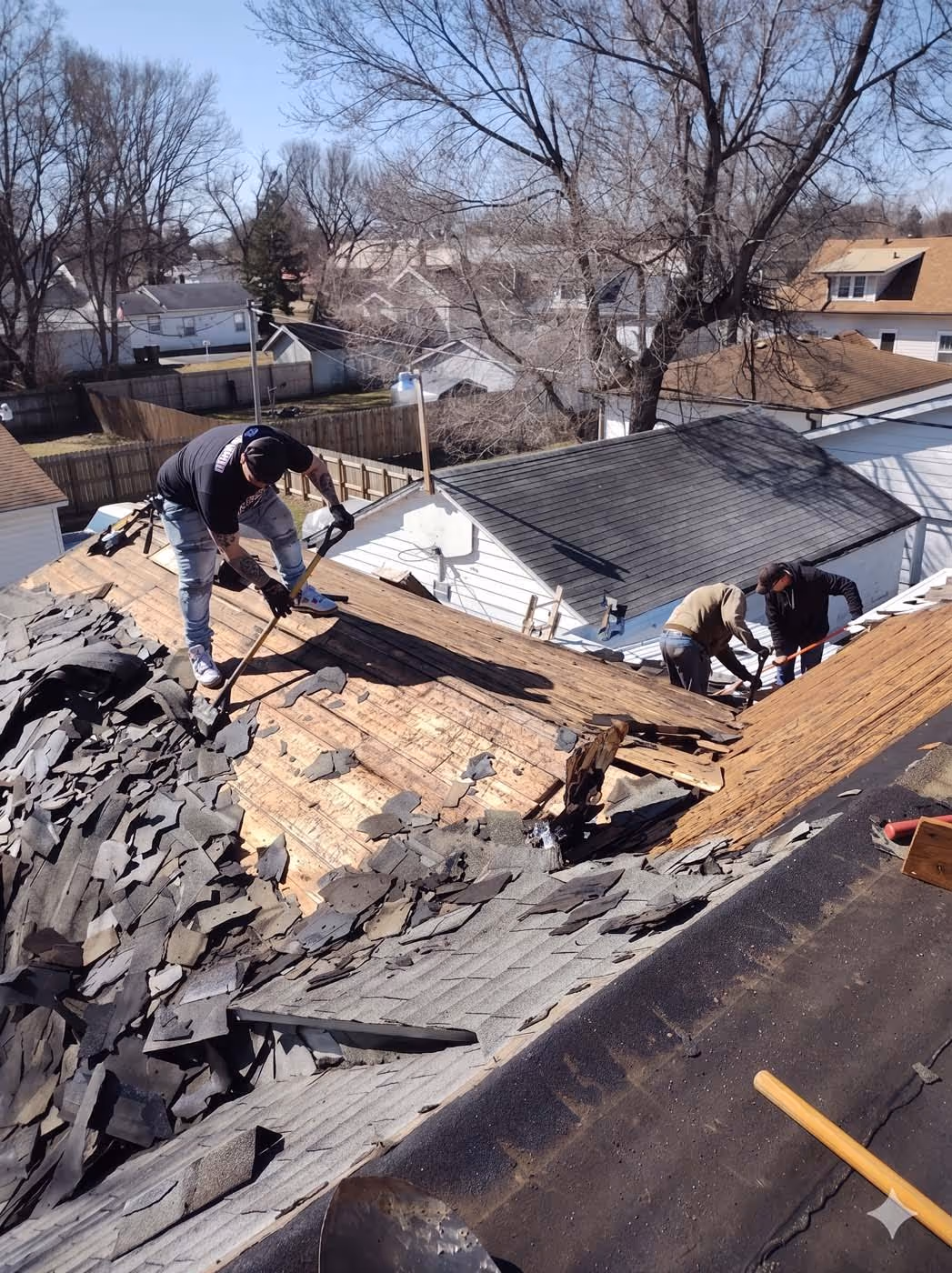 Three workers removing old shingles from a residential roof under clear sky with leafless trees in the background.