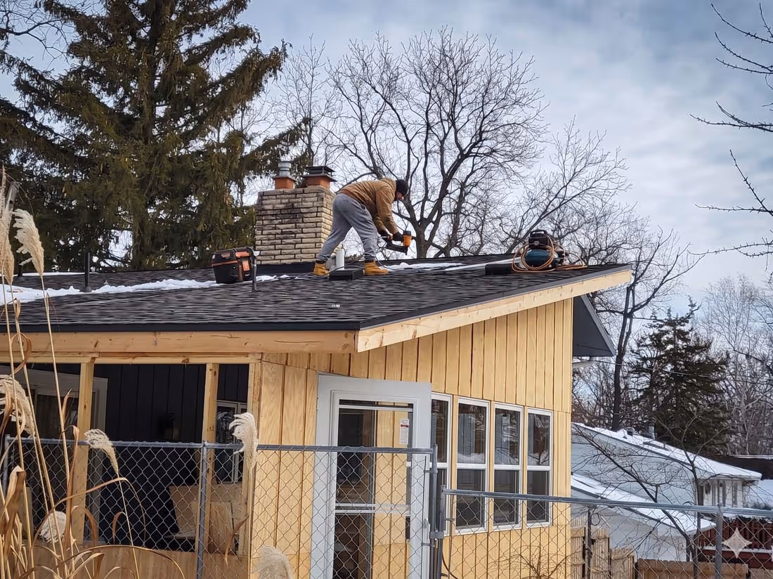 Worker installing black shingles on the roof of a wooden house during winter with bare trees in the background.