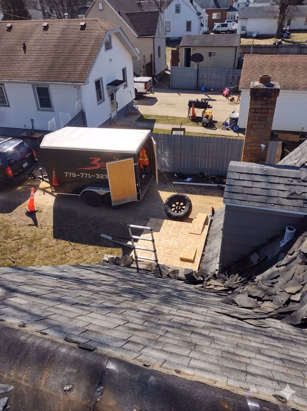 View from a rooftop showing worn shingles and a work area below with a trailer, ladder, spare tire, and residential houses.