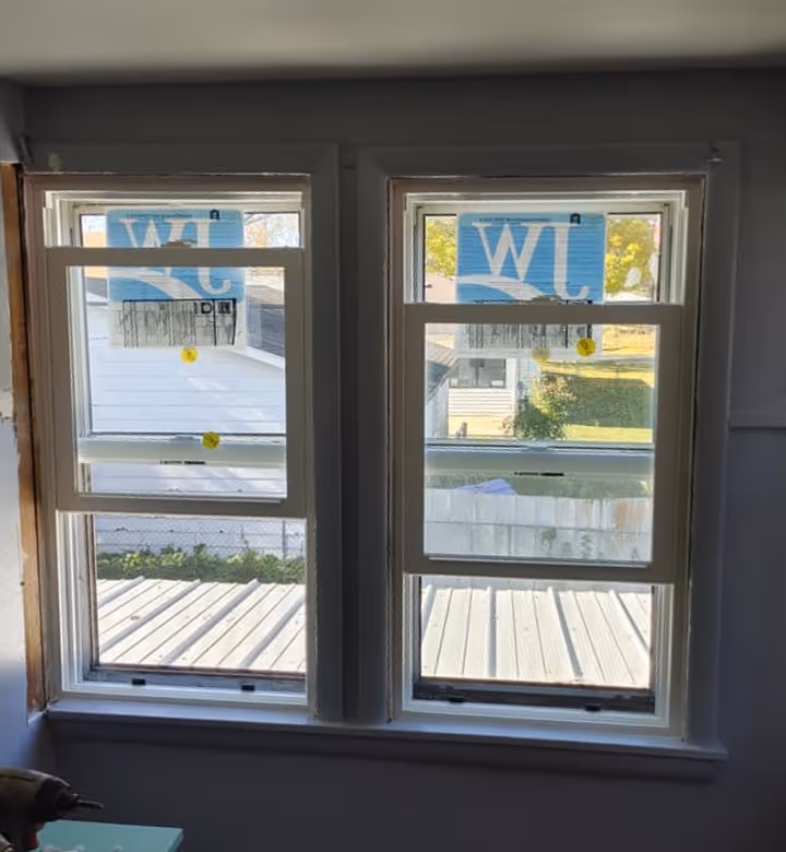 Two white-framed double-hung windows installed side by side with blue manufacturer labels still attached.
